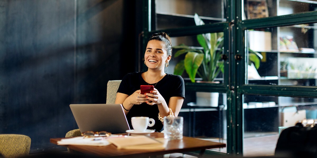 woman sitting at table holding phone, laptop and coffee cup on the table