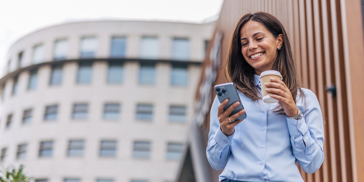 professional woman smiling, standing outside of building, holding cup of coffee and cellphone